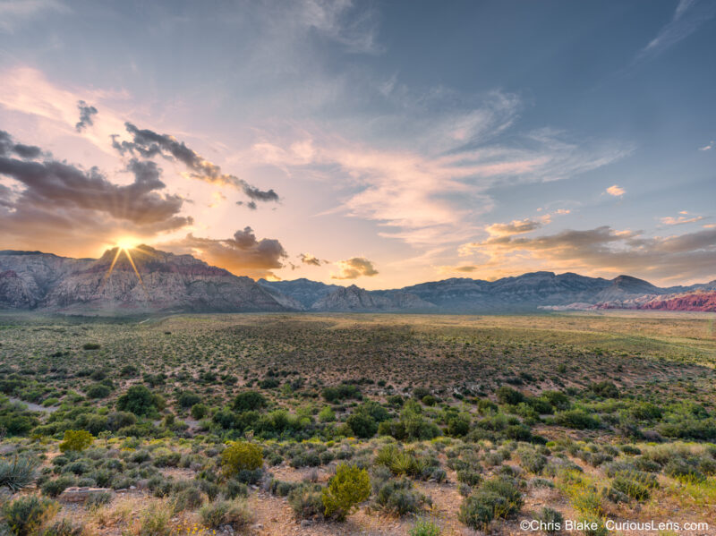 Sunset at Red Rock Canyon near Las Vegas, showcasing the vibrant colors of the sun as it sets behind the mountain range. This panoramic view captures the fading light across the valley, with a brilliant sunstar and a rich palette of red and gold tones. The image conveys the tranquility and natural beauty found just outside the bustling Las Vegas Strip.