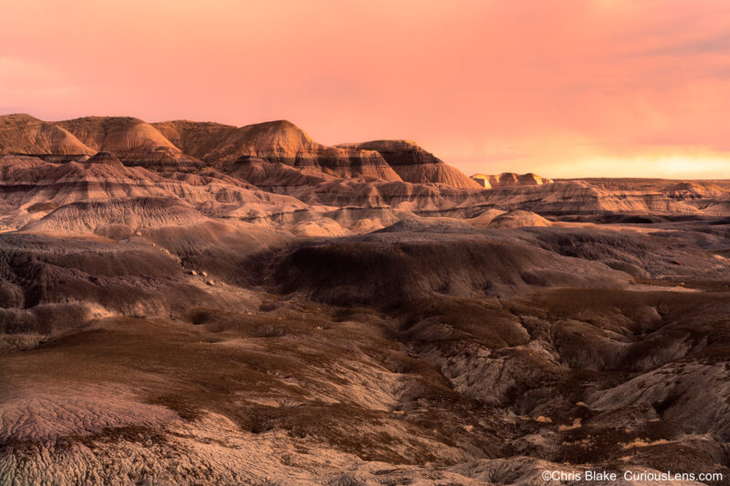 Sunset view from the Blue Forest Trail in Petrified Forest National Park. This historic trail, first constructed in 1934 and reopened in 2013, offers challenging terrain through badlands hills. On this evening hike, the setting sun illuminated distant storm clouds in vibrant red, casting a warm glow across the landscape. This photo captures the dramatic contrast between the blue badlands and the sunset's red hues, showcasing the trail's stunning and ever-changing scenery.