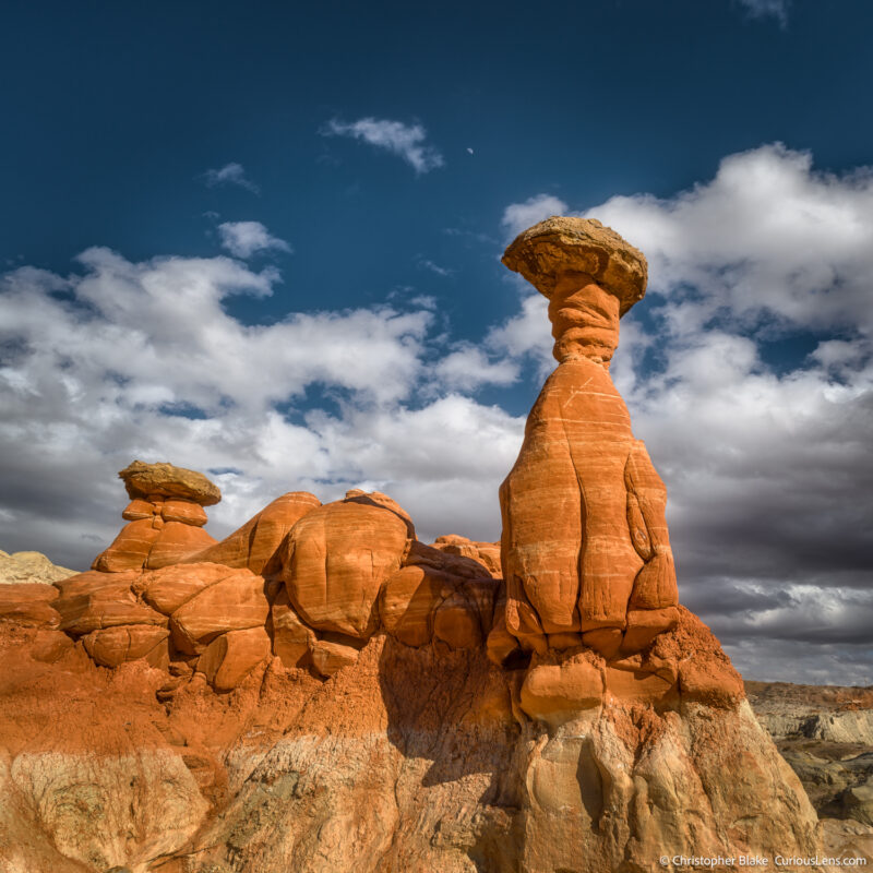 The Toadstool Hoodoos in the Grand Staircase-Escalante National Monument feature striking rock formations formed over millions of years. This photo captures a tall hoodoo with a smaller one beside it, framed by dramatic sandstone formations and a darkening sky with storm clouds. The image was taken in the late afternoon, emphasizing the textures and unique shapes, with the angle from below accentuating the hoodoos against the blue sky. The scene conveys the area's fragile beauty and the need for careful exploration.