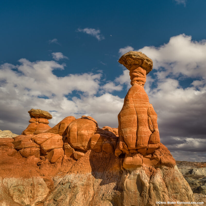 The Toadstool Hoodoos in the Grand Staircase-Escalante National Monument feature striking rock formations formed over millions of years. This photo captures a tall hoodoo with a smaller one beside it, framed by dramatic sandstone formations and a darkening sky with storm clouds. The image was taken in the late afternoon, emphasizing the textures and unique shapes, with the angle from below accentuating the hoodoos against the blue sky. The scene conveys the area's fragile beauty and the need for careful exploration.