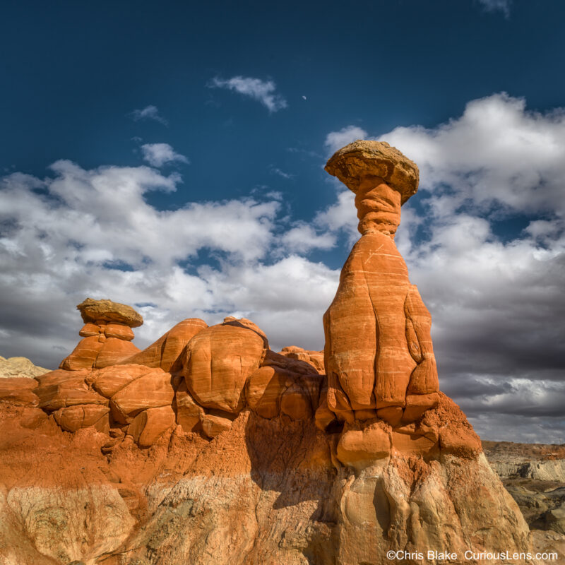 The Toadstool Hoodoos in the Grand Staircase-Escalante National Monument feature striking rock formations formed over millions of years. This photo captures a tall hoodoo with a smaller one beside it, framed by dramatic sandstone formations and a darkening sky with storm clouds. The image was taken in the late afternoon, emphasizing the textures and unique shapes, with the angle from below accentuating the hoodoos against the blue sky. The scene conveys the area's fragile beauty and the need for careful exploration.