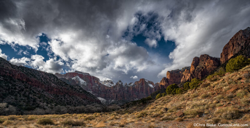 A panoramic view of the Towers of the Virgin in Zion National Park, captured in the late afternoon. The scene includes the West Temple, the Sundial, and the Altar of Sacrifice, with clouds breaking over the peaks and fresh snow visible. The lighting varies across the valley, with some areas in shadow and others illuminated by the sun. This image was taken as a winter storm was dissipating, showcasing the dramatic contrast between light and shadow in this iconic landscape.