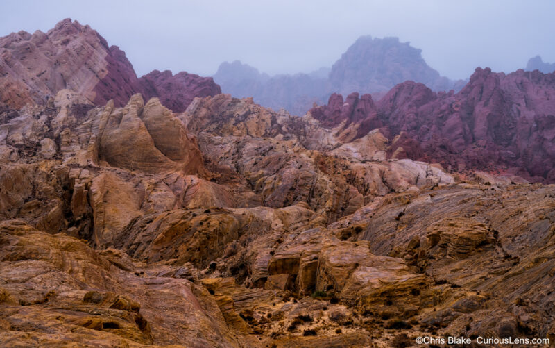 Photograph taken in Valley of Fire during a rare rainy and foggy early winter morning. The scene captures a dramatic landscape with rocks changing in color and shape from the foreground to the background, with the distant formations fading into a misty horizon. The soft rain and fog create an unusual and atmospheric setting, providing a unique perspective on the typically arid landscape of southern Nevada.