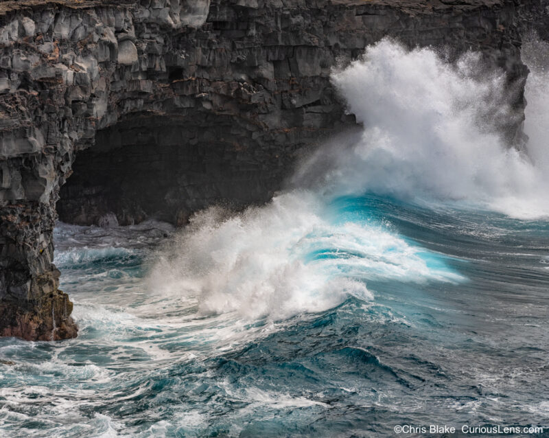 Massive ocean wave crashes into volcanic cliffs and a lava tube along the Chain of Craters Road, preserving the intricate details of water and mist against the rugged terrain.