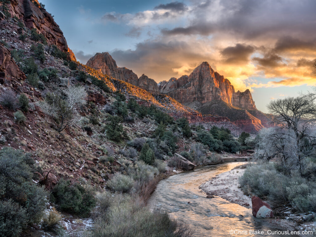 A dramatic sunset over the Watchman at Canyon Junction Bridge in Zion National Park. The river leads toward the towering mountain peaks in the distance, with a vibrant red hue lighting up the clouds as the sun sets. The scene captures a blend of natural beauty, with the last light of the day highlighting the landscape and reflecting off the river's surface. The composition of the image emphasizes the convergence of water and mountains, creating a stunning, picturesque view.