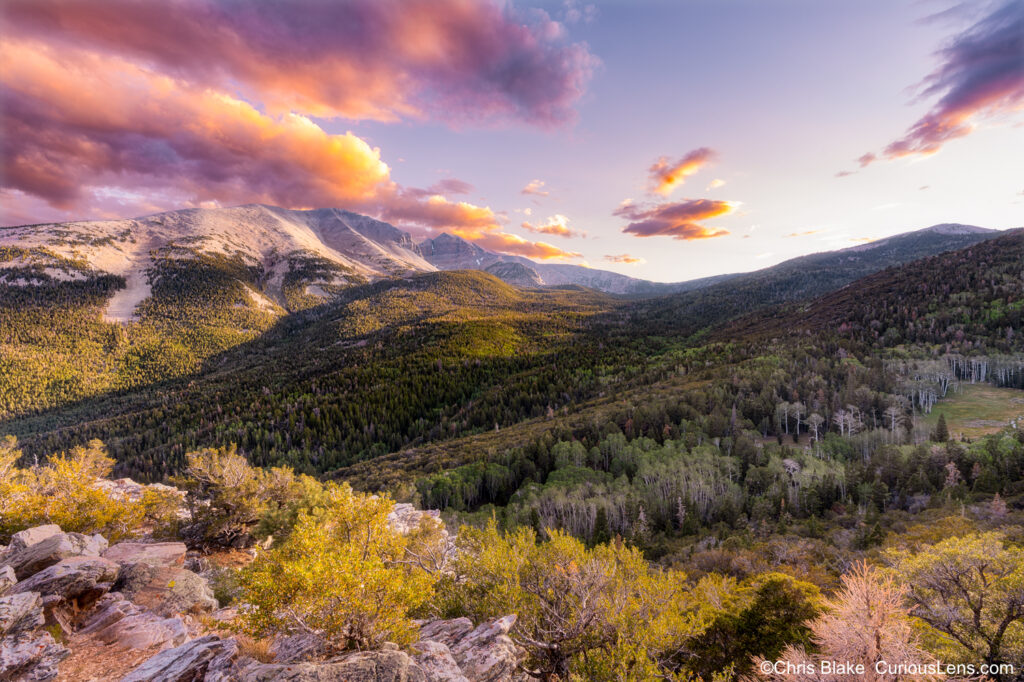 Great Basin National Park at sunset, showing Wheeler Peak and colorful clouds over a pine and aspen-covered valley, captured from a rocky mountainside at 9,000 feet elevation."