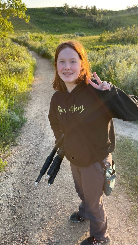 Cameron making a peace sign while the soft sun lights up her red hair, carrying my tripod down a trail in Theodore Roosevelt National Park.