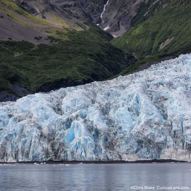 "Aialik Glacier in Aialik Bay, Kenai Fjords National Park, with serene waters, jagged ice, towering mountains, sunlight on peaks, and cascading waterfalls.