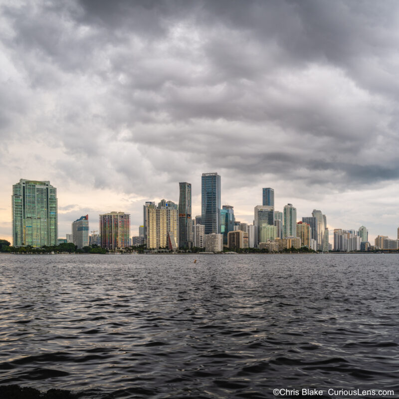 Thunderstorm over Miami captured from Rickenbacker Causeway with lightning, brooding clouds, sunlight on buildings, and tranquil bay waters.