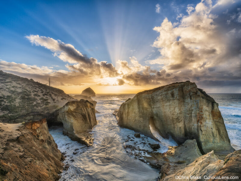 Cape Kiwanda, Oregon at sunset with sun rays through clouds, rain over the ocean, storm clouds, crashing waves, and glowing arch.