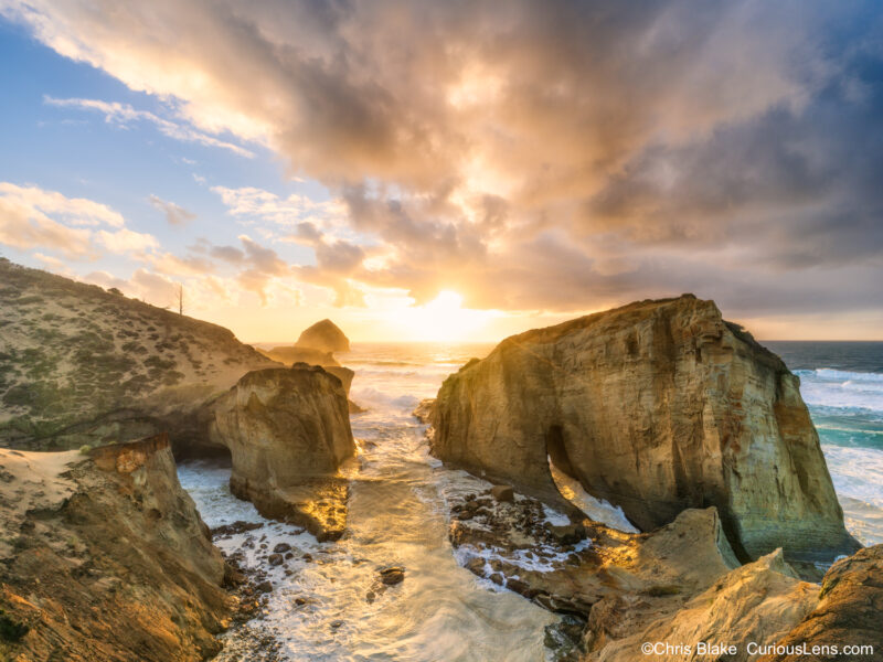 Cape Kiwanda on the central Oregon coast at sunset with golden glow on water and rocks, fiery red storm clouds, and massive waves crashing in the distance.