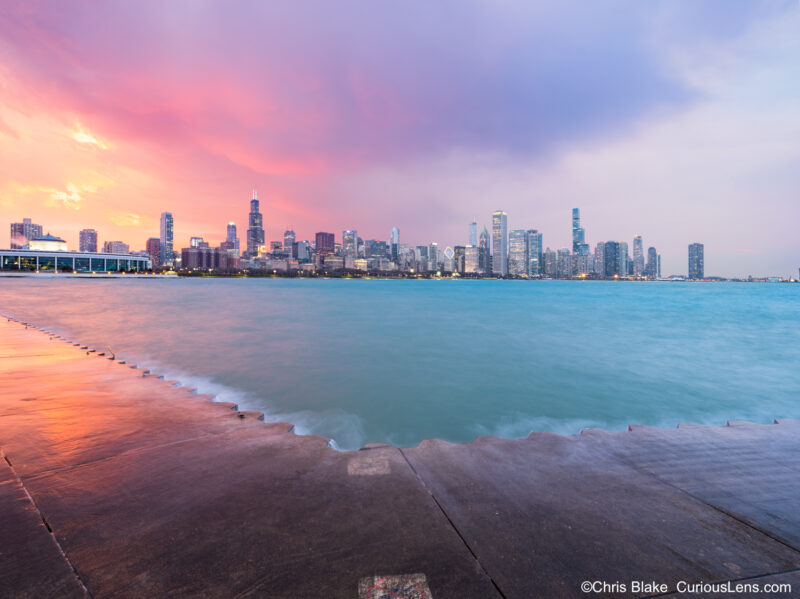 Chicago skyline at sunset from Skyline Walk with Willis Tower, Aon Center, John Hancock Center, storm clouds, building lights, and choppy lake reflections.