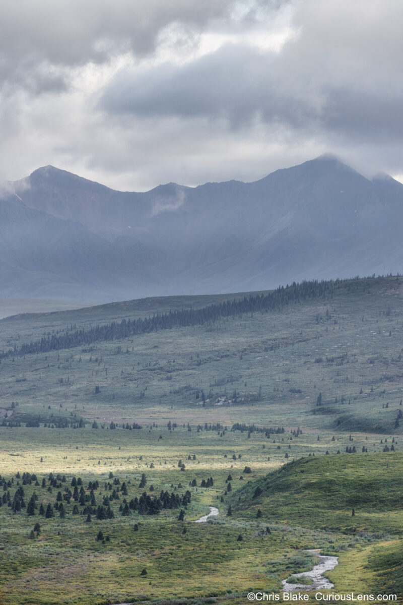 Serpentine river meandering through emerald hills with mist-draped majestic mountains in the distance, captured before a storm in the valley."