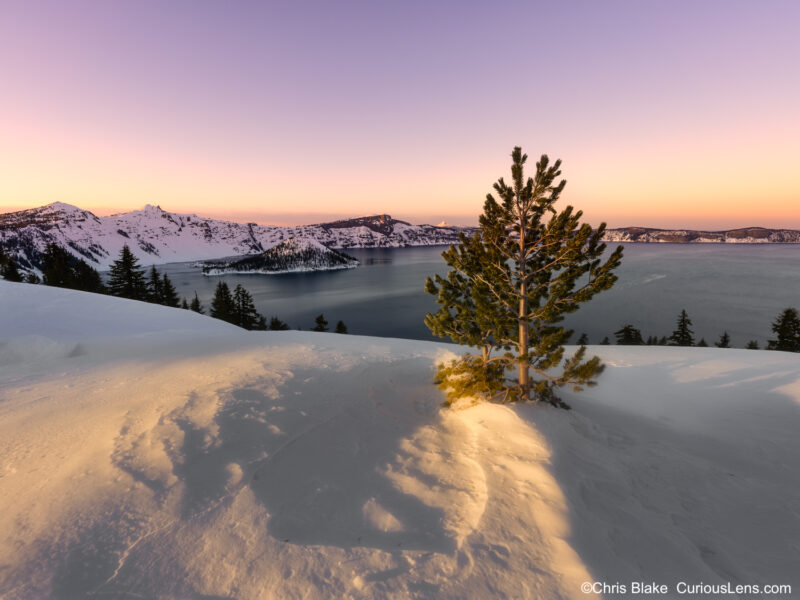 Winter at Crater Lake National Park with snow-covered crater, warm pink and red sunset sky, and deep blue water.