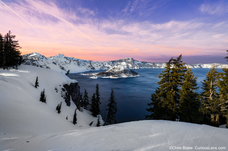 Winter at Crater Lake National Park with snow-covered crater, warm pink and red sunset sky, and deep blue water.