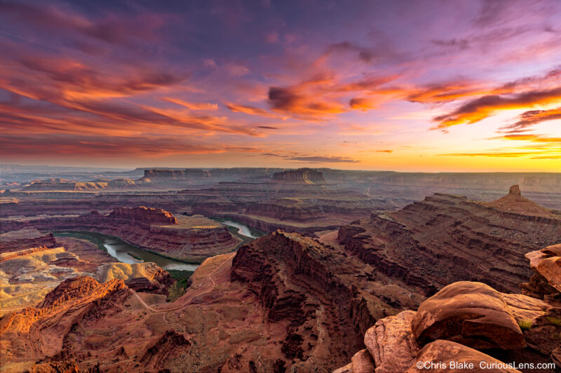 Sunset at Dead Horse Point State Park with gooseneck in Colorado River, vertical cliffs, colorful rocks, and vibrant red clouds.