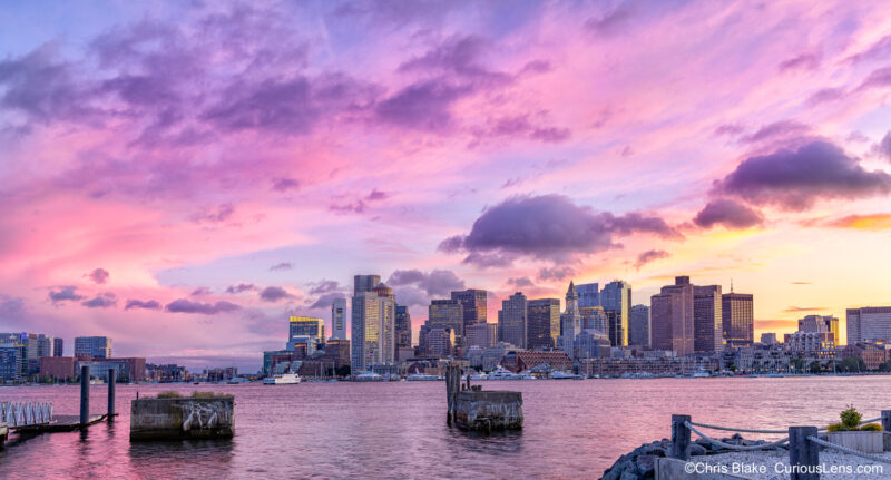 Sunset over downtown Boston from East Boston with pink and red clouds, bay, old pier remains, Long Wharf, and Custom House.