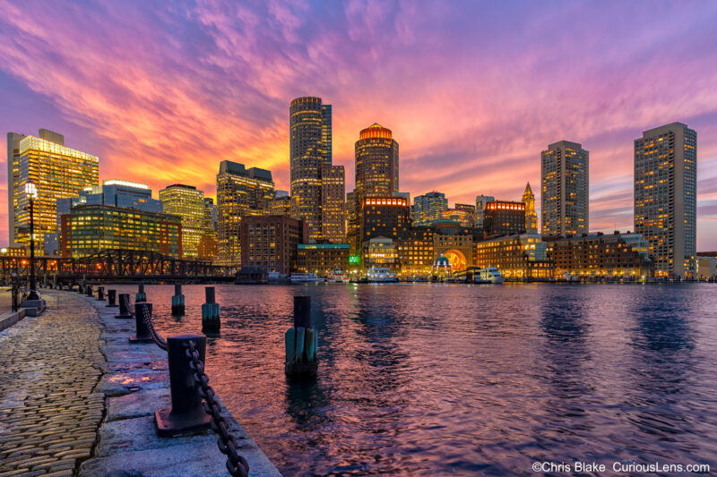 Winter sunset over downtown Boston from Fan Pier Park with vibrant clouds, reflections in the bay, and city lights.