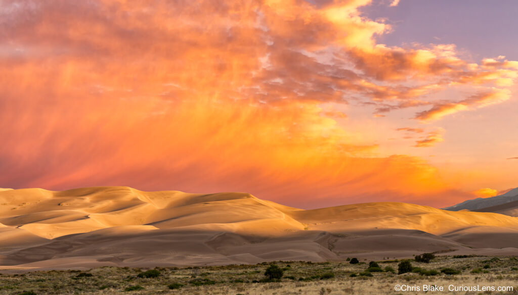 Sunrise over Great Sand Dunes with red storm clouds, soft morning light, and Sangre de Cristo Mountain Range for scale.