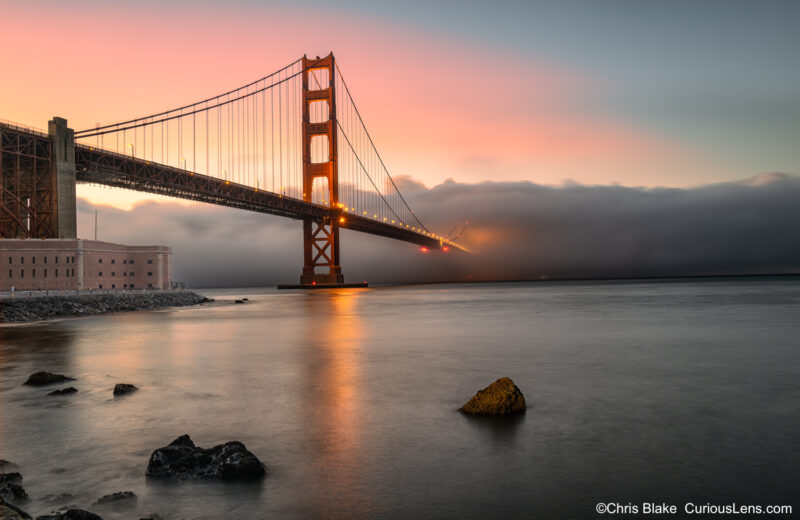 Fort Point and Golden Gate Bridge with fog covering half the bridge, sunset colors in the sky, and unique water perspective.