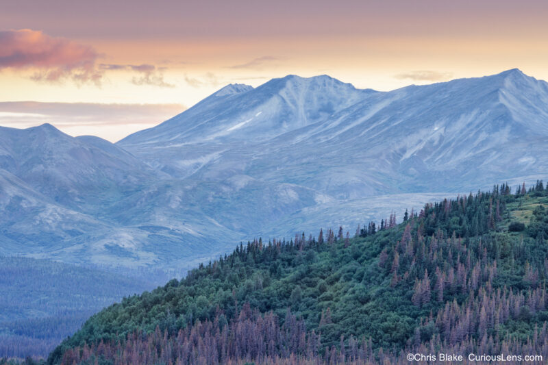 Sunset view from George Parks Highway in Denali National Park with vibrant sky, sharply defined foreground mountain, and distant mountains softened by haze and smoke.