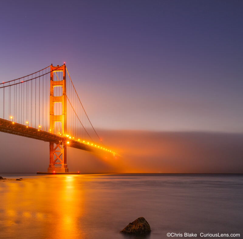 Golden Gate Bridge during blue hour with fog, long exposure soft water, vibrant orange bridge, and empty sky.