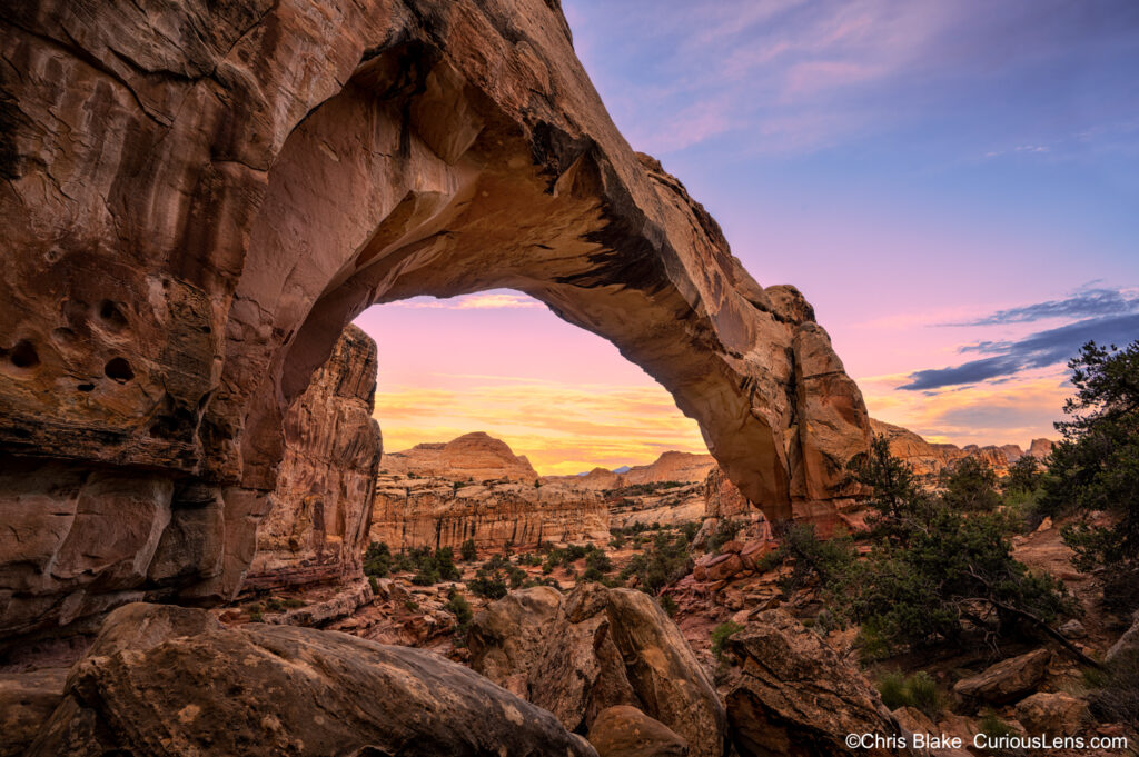 Sunset at Hickman Natural Bridge in Capitol Reef with colorful clouds, valley view, boulders in foreground, and dramatic arch.