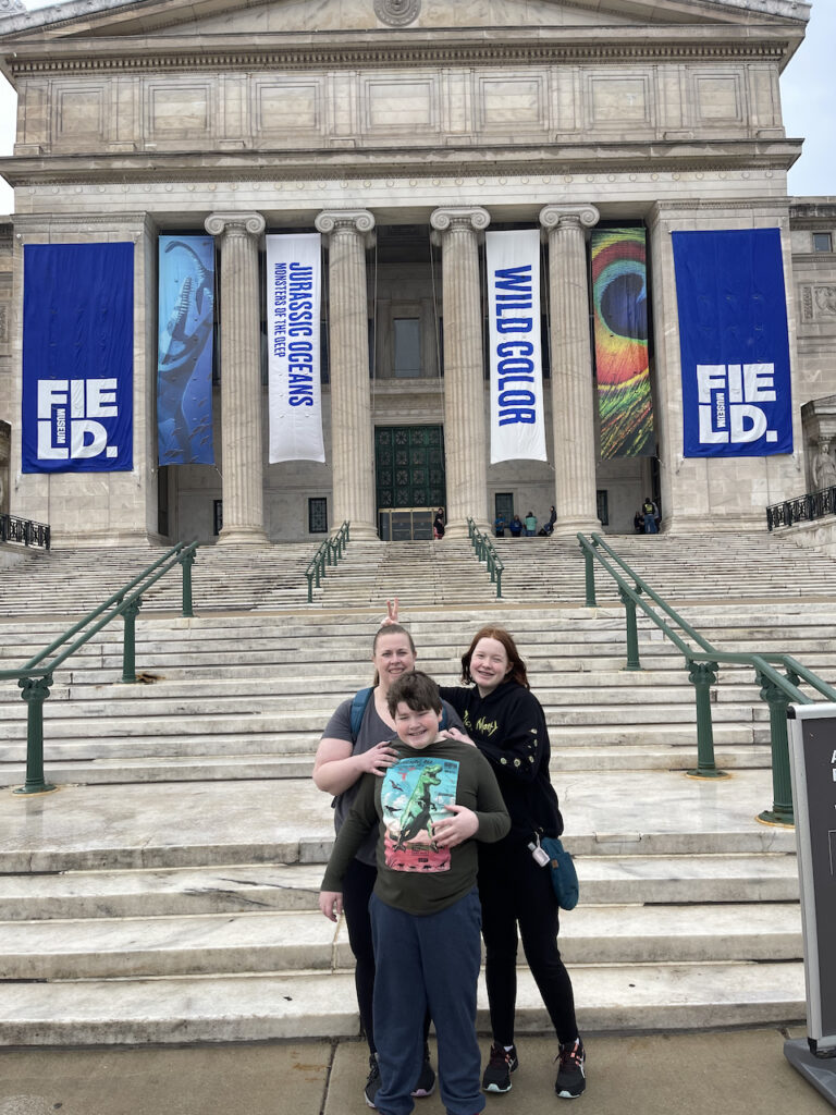 Cat, Collin and Cameron at the front steps of the Field Museum, everyone is smiling and Cameron is giving Collin Bunny ears.