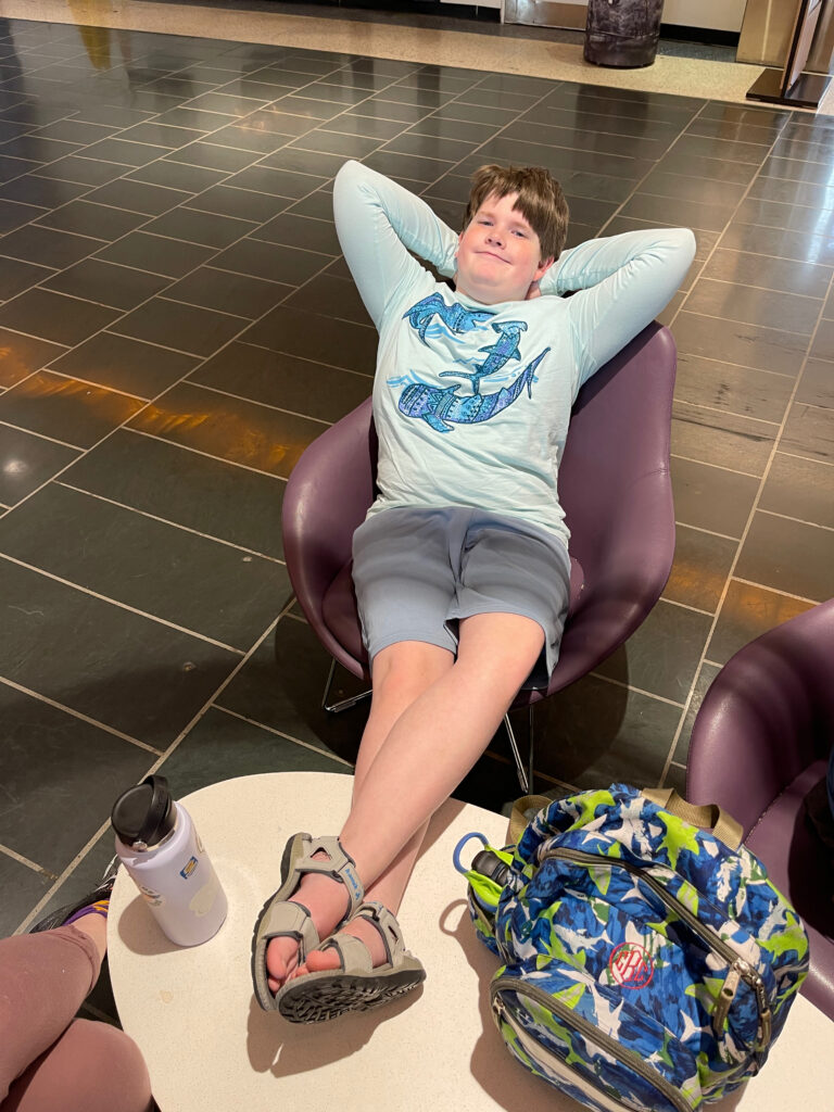 Collin sitting back in a purple chair with his hands behind his head and his feet up on a table in Boston Logan airport.