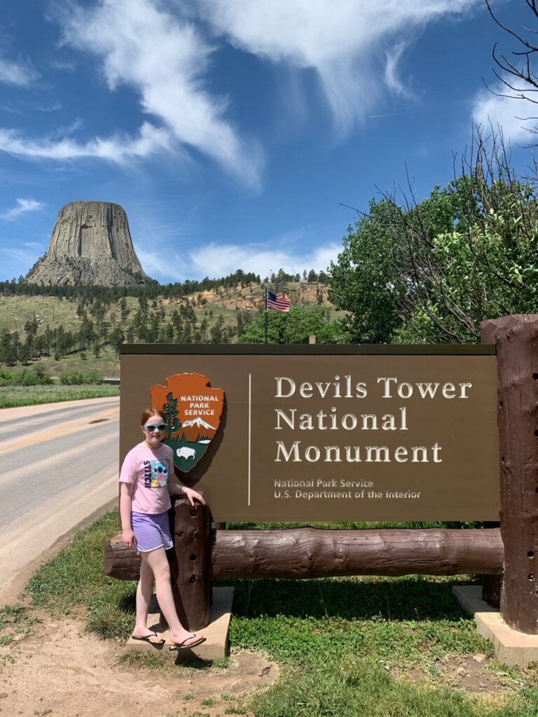 Cameron standing in front of the Devils Tower National Monument park sign. The tower is in the background with a deep blue sky full of clouds. 