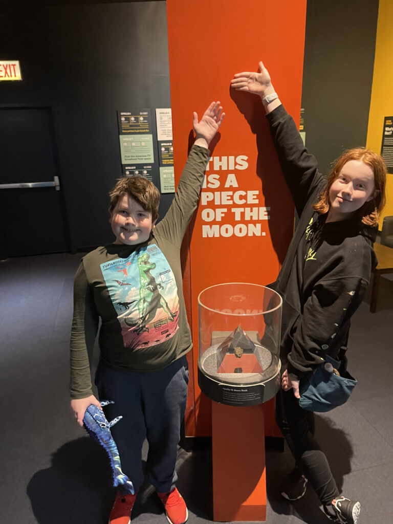 Cameron and Collin with one arm up each, showing off a piece of the moon in the Adler Planetarium.