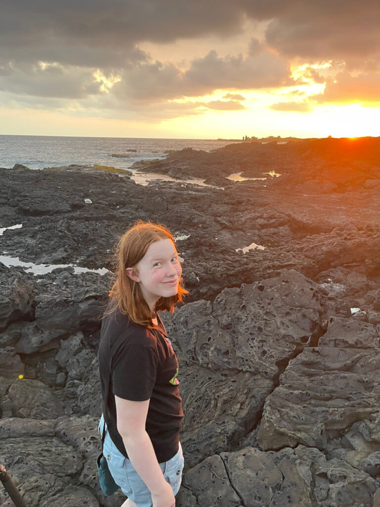 Cameron standing on lava rack at Beans Beach at sunset as the golden light makes her red hair glow. The Pacific ocean is calm as we watch the clouds above light up.