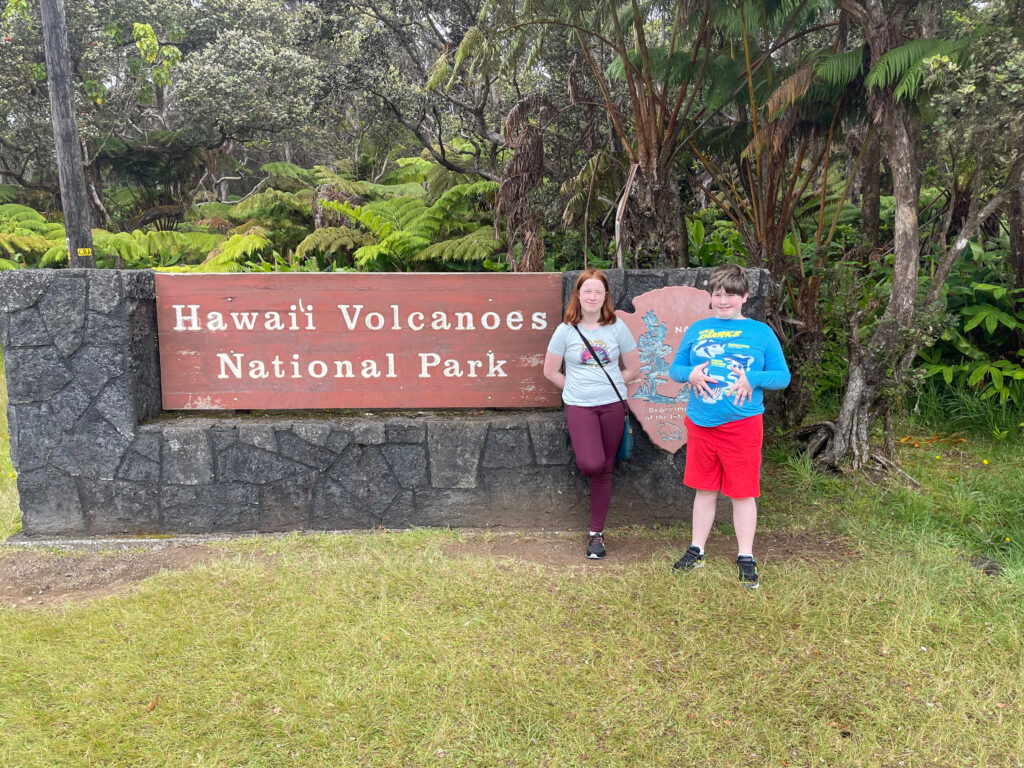 Cameron and Collin standing in front of the Hawaii Volcanoes National Park sign. Both all smiles with the jungle behind them. 