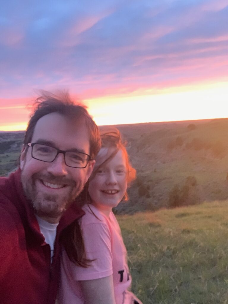 Cameron and myself posing at sunset, with a red sky, and warm red last cast over us. At the Biocourt Overlook in Theodore Roosevelt National Park.