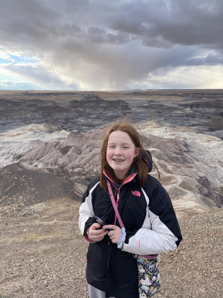 Cameron holding her phone on top of the tallest badlands on the Blue Forest Trail. You can see storm clouds and rain in the distance and an epic valley below.