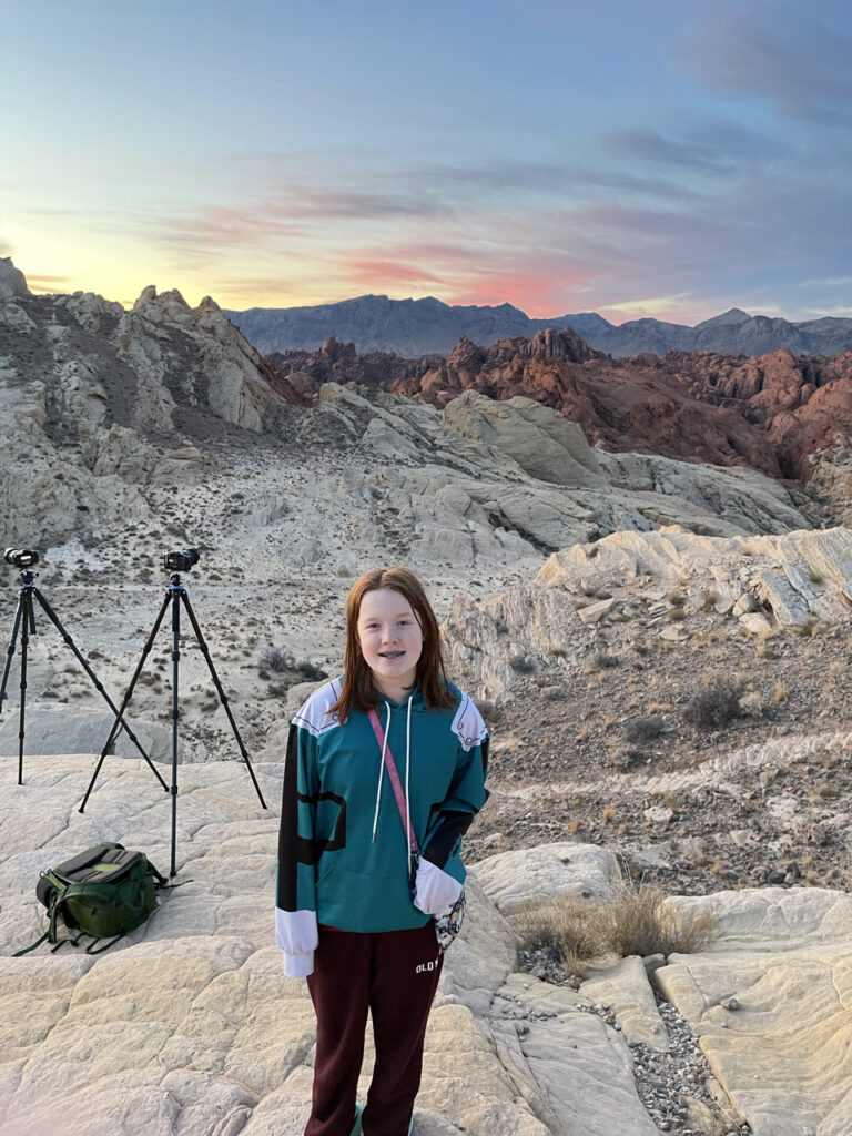 Cameron standing on the rocks at sunset near the Silica Dome in the Valley of Fire State Park. The sky is full of red and yellow clouds and the red rocks in the background are almost glowing. My tripods can be seen in the background.