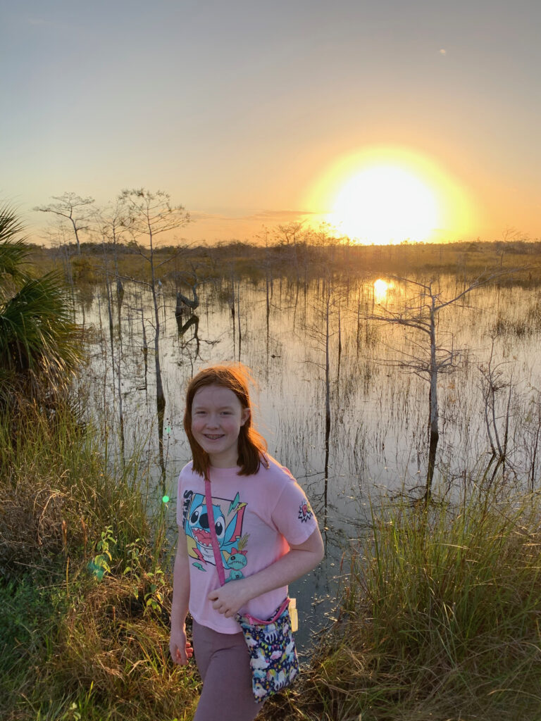 Cameron at sunrise at the shores of a lake in the Everglades National Park. The sky is a bright yellow and one of the famous Z-trees can be seen in the water behind her.