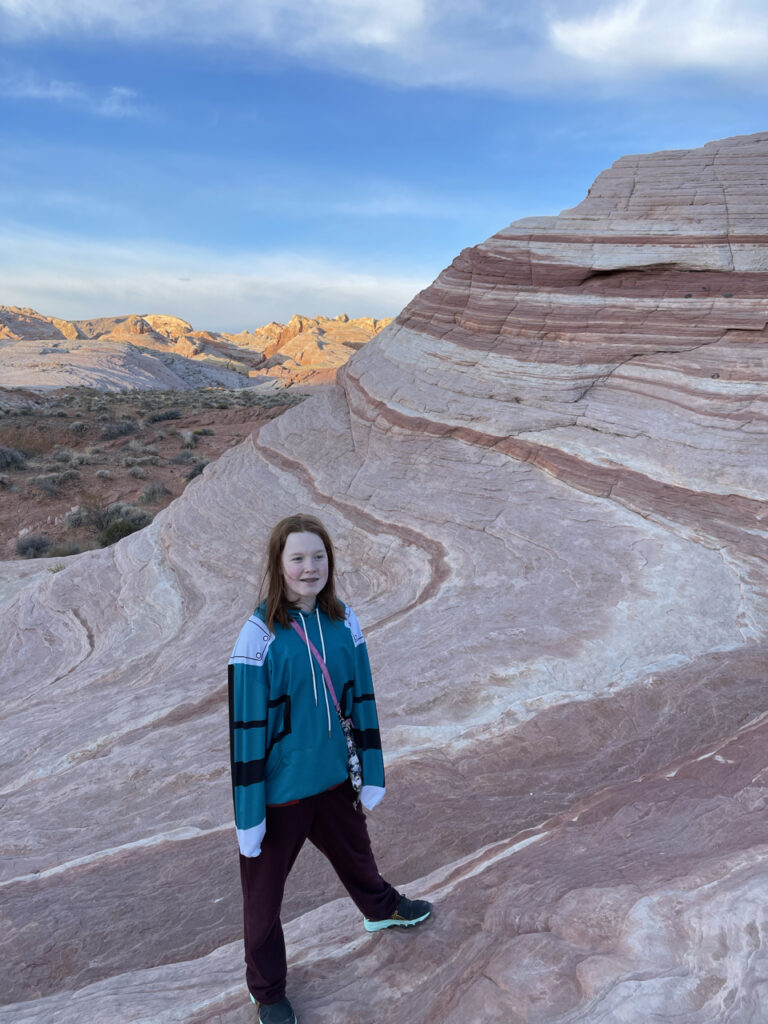 Cameron at sunrise at the Fire Wave in the valley of Fire State park. She is standing halfway up the main "wave" or hill with a big smile on her face and the sunlight has just stated to reach the mountain range in the far background.