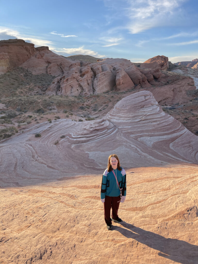 Cameron standing on the Fire Wave in the Valley of Fire State Park. The sun has just come up and is casting warm yellow light over her, but the background of the wave is still in shadows.