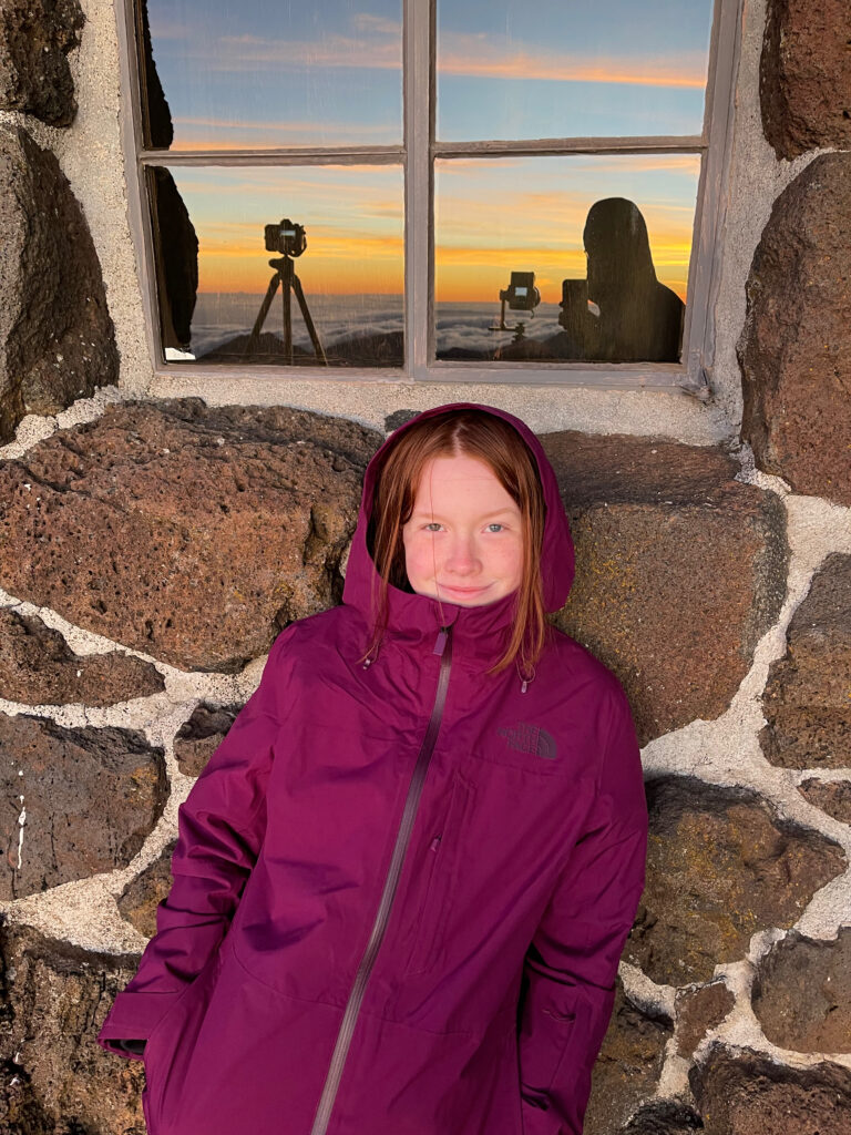 Cameron in her purple winter coat leaning on the brick visitors center wall at Haleakala National Park. The window above her has a perfect reflection of the sunrise she is looking at, with a full cloud inversion and yellow and red clouds. 