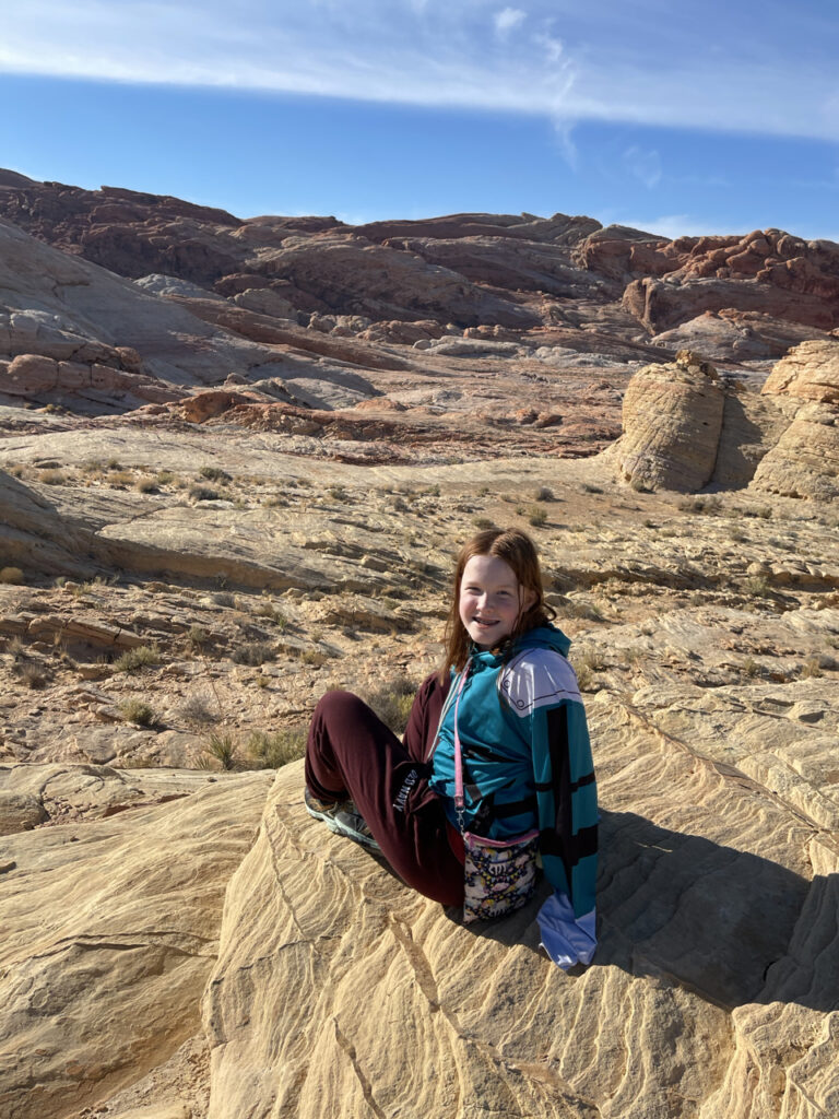 Cameron sitting on a giant rock ledge at a random view point we climbed too in the Valley of Fire State Park. She is smiling and happy with the amazing view of the valley and mountains. 