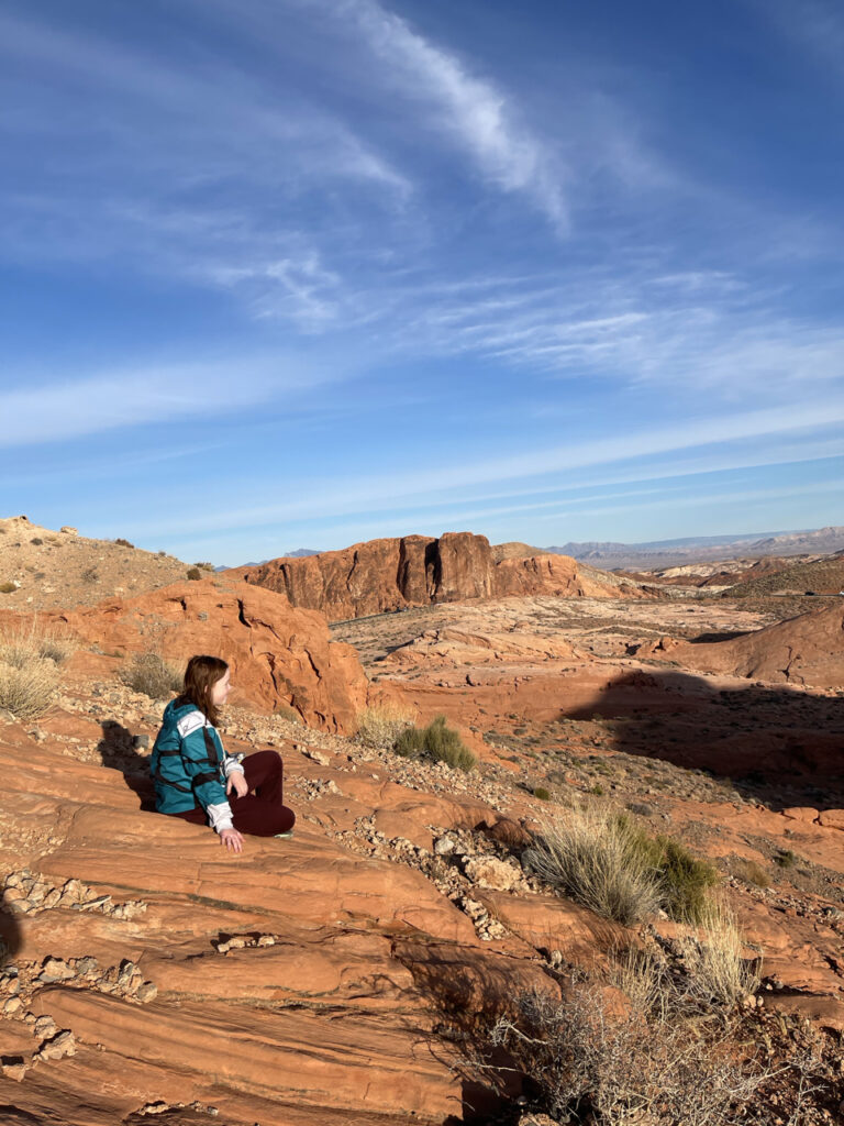 Cameron sitting done on the red rocks near the top of the mountain at the White Domes trail. The shadows are growing long in the frame as the sun gets lower and the sky has some wispy clouds.