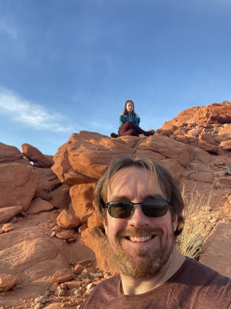 Me standing on a trail in the Valley of Fire State Park, with the soft yellow sunset light on my face. Above me is Cameron sitting on the rocks that are also awash with the warm glowing light.