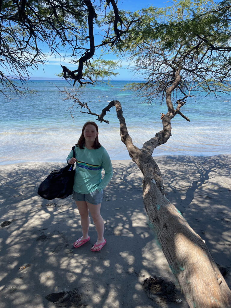 Cameron standing in the shake on Olowalu Beach just feet from the ocean holing a beach bag.A