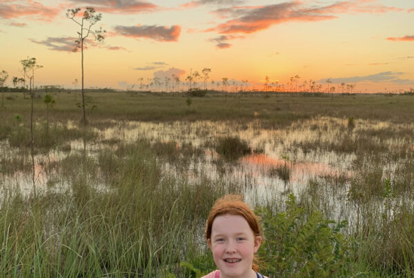 Cameron in the Everglades smiling at sunset as the sky lights up with color and it is reflected in the swamp behind her.