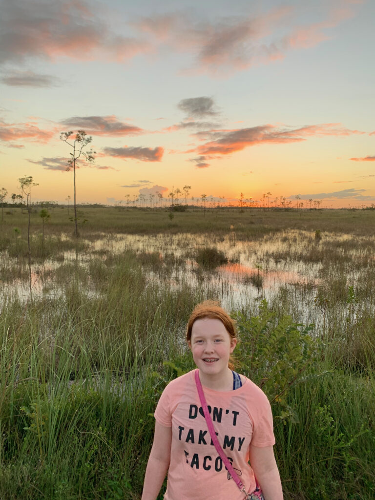 Cameron in the Everglades smiling at sunset as the sky lights up with color and it is reflected in the swamp behind her.