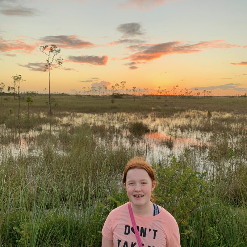 Cameron in the Everglades smiling at sunset as the sky lights up with color and it is reflected in the swamp behind her.