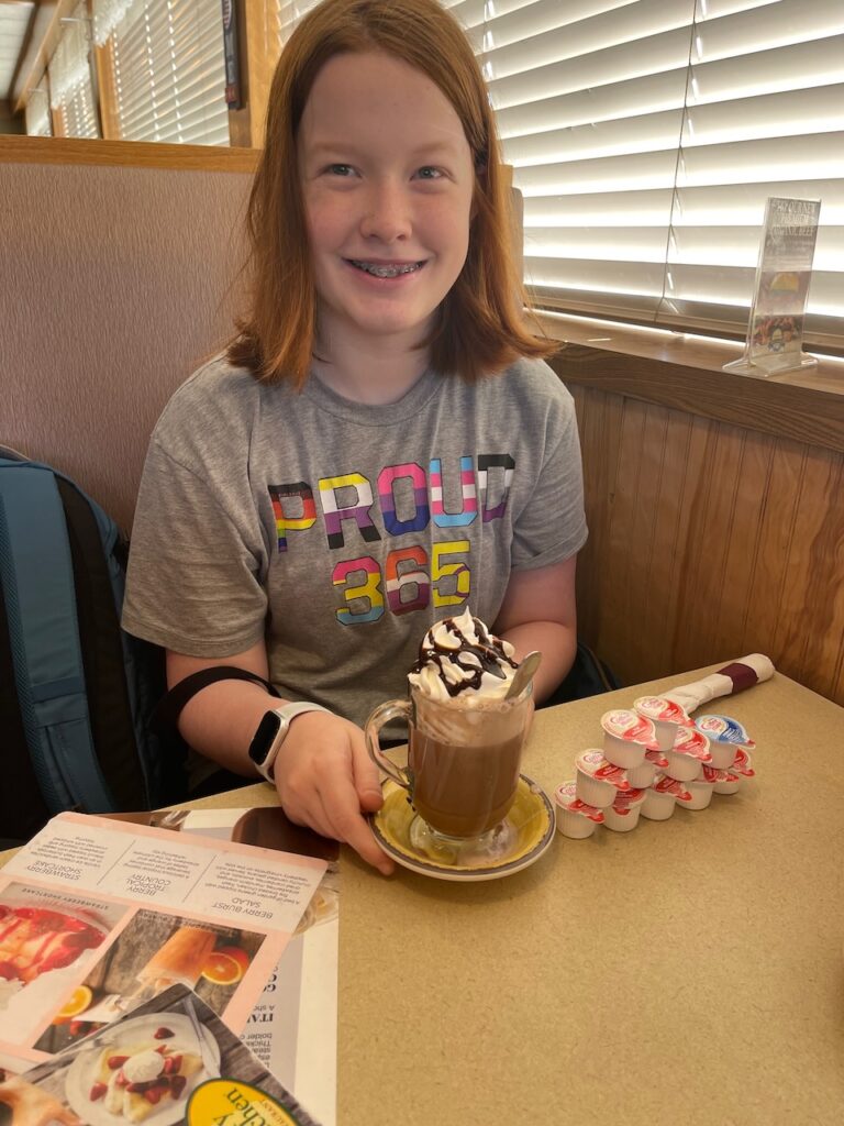 Cameron with her red hair and pride shirt sitting down for hot chocolate at a restaurant outside of Theodore Roosevelt National Park. 