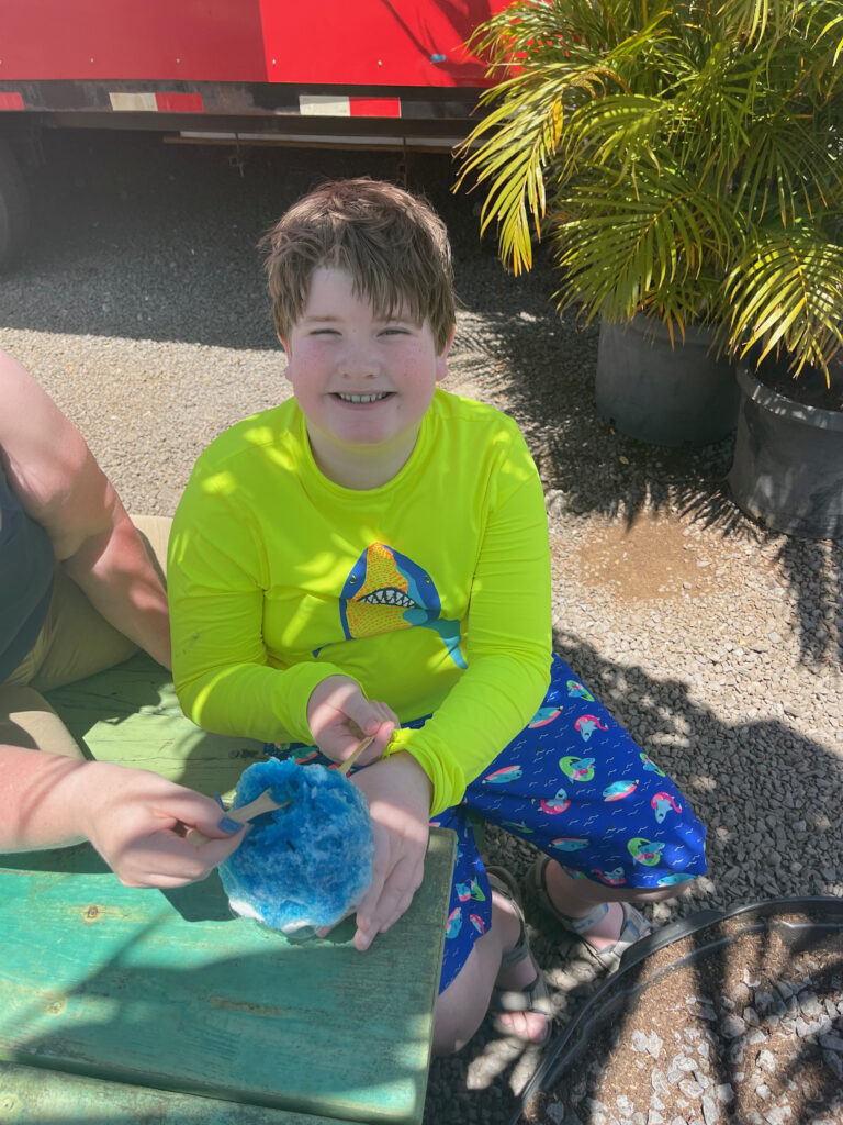 Collin sitting at an outdoor table, with a food truck behind him eating frozen ice in Maui Hawaii. 