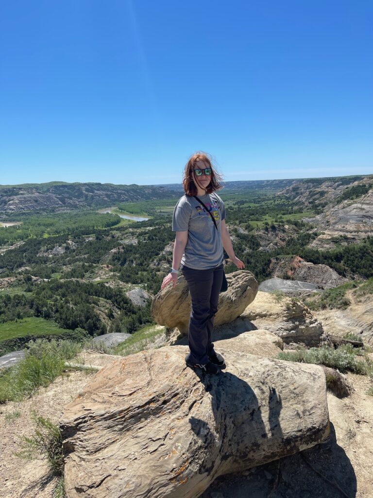 Cameron standing on top of a pile of massive rocks, in the Badlands of Theodore Roosevelt National Park. The green valley and Little Missouri River are in the background.